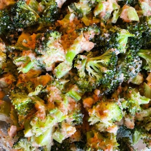 Close-up of cooked broccoli florets coated in a creamy, orange-hued sauce, with visible seasoning and small bits of ingredients mixed throughout.