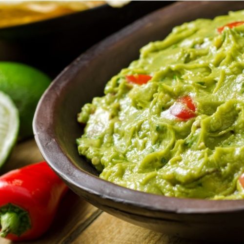 A close-up of a bowl of guacamole garnished with tomato pieces and cilantro. A lime wedge and a red chili pepper are on the wooden table beside the bowl.