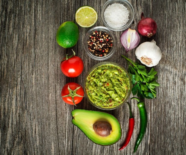 A bowl of guacamole surrounded by fresh ingredients: avocado, tomatoes, lime, red onion, garlic, parsley, chili peppers, salt, and peppercorns, all arranged on a rustic wooden surface.