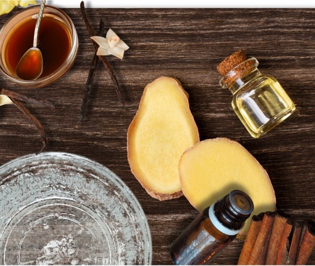 Sliced ginger, cinnamon sticks, vanilla beans, a small glass jar of oil, a brown bottle, and a bowl of honey with a spoon arranged on a rustic wooden surface.