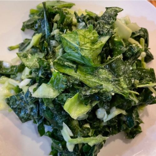 A close-up of a plate of chopped kale salad coated in a creamy dressing, served on a white plate atop a wooden surface.