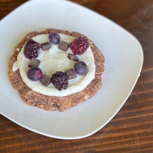 A round cookie topped with white frosting, blackberries, blueberries, and small chocolate chunks sits on a white plate on a wooden table.