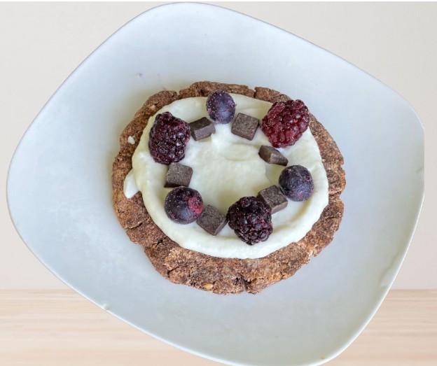 A round, brown cookie topped with white yogurt, blackberries, and small chocolate cubes sits on a white, slightly tilted plate against a light background.