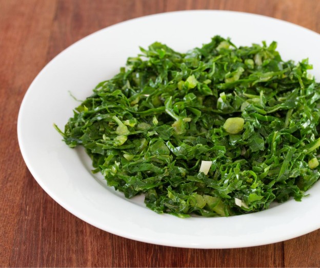 A white plate filled with finely chopped collard greens sits on a wooden table. The greens appear fresh and vibrant, with a glossy texture.