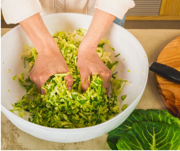 A person’s hands mixing or squeezing shredded cabbage and greens in a large white bowl, with a cutting board, knife, and cabbage leaves nearby on the counter.