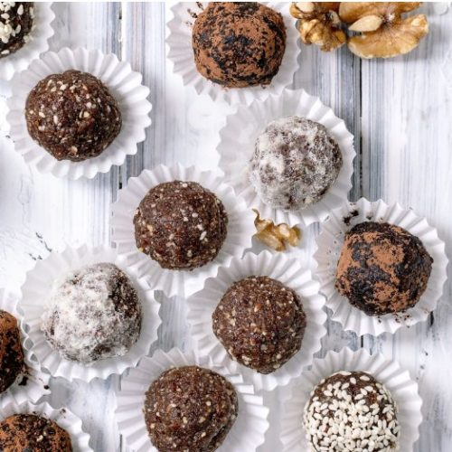 Assorted chocolate truffles dusted with cocoa, powdered sugar, and sesame seeds, arranged in white paper cups on a white wooden surface, with walnut pieces scattered nearby.
