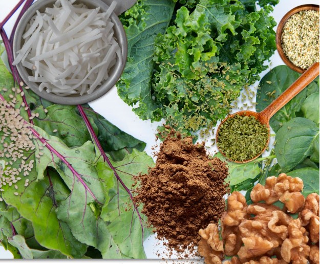 An assortment of healthy foods including kale, spinach, beet greens, walnuts, dry noodles, ground spices, sesame seeds, and dried herbs on a white background.