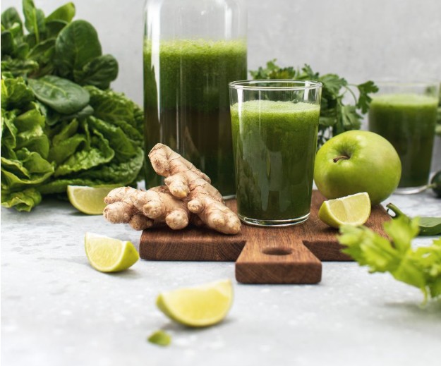 A glass and bottle of green juice on a wooden board, surrounded by fresh spinach, lettuce, ginger root, lime wedges, a green apple, and parsley on a light surface.