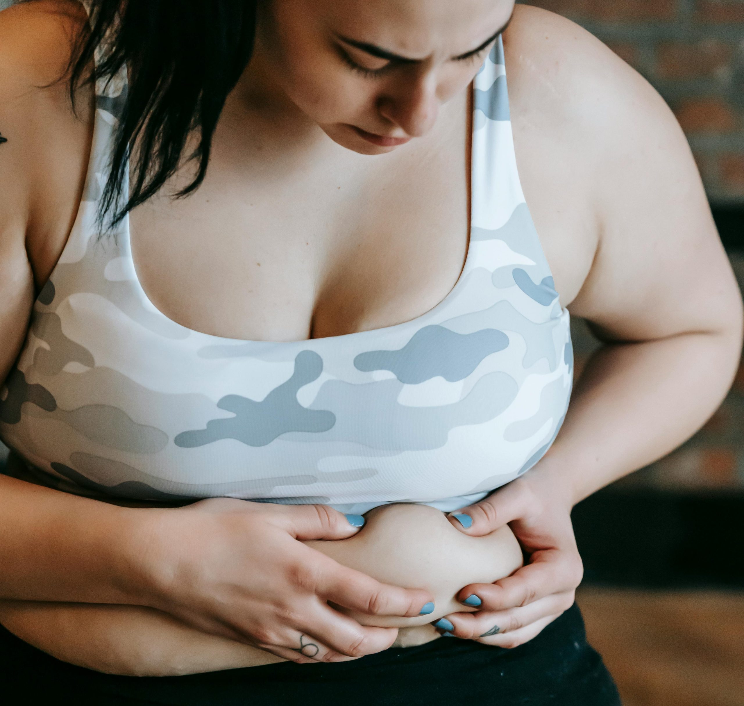 A woman in a camouflage sports bra looks down and gently pinches her stomach with both hands, showing body positivity.