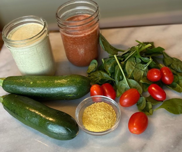 Two glass jars with smoothies, two zucchinis, grape tomatoes, fresh spinach, and a small bowl of nutritional yeast are arranged on a marble surface.