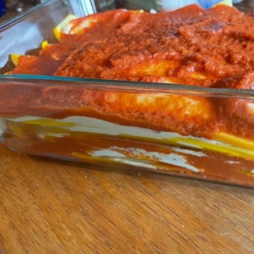 A close-up of an unbaked glass dish of lasagna, showing visible layers of pasta, creamy white cheese, and red tomato sauce on a wooden surface.