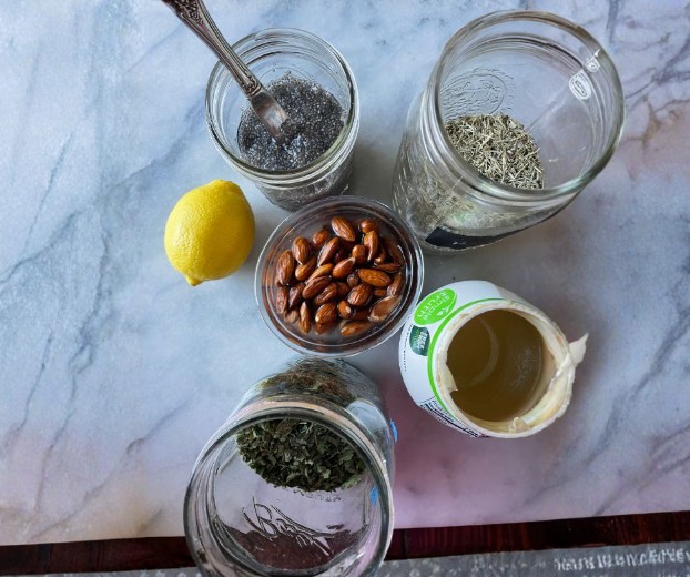 Five jars containing chia seeds, rosemary, dried herbs, almonds, and coconut oil are arranged on a marble surface. A metal spoon is in one jar, and a whole lemon sits beside the jars.