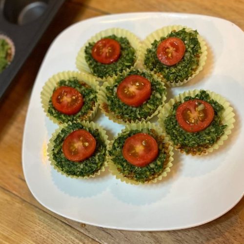 Seven mini savory muffins topped with halved cherry tomatoes are arranged on a white plate. The muffins appear to contain spinach or herbs and are in yellow paper cups, set on a wooden table.