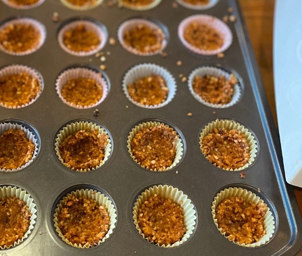 A muffin tin filled with paper liners, each holding a portion of a crumbly baked mixture, possibly homemade granola or muffin bases. The tin is resting on a wooden surface.