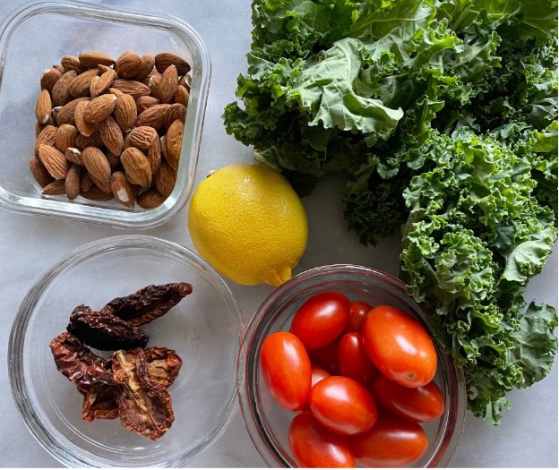 A top view of fresh kale leaves, a lemon, grape tomatoes, whole almonds in a glass container, and sun-dried tomatoes in a small bowl on a light surface.