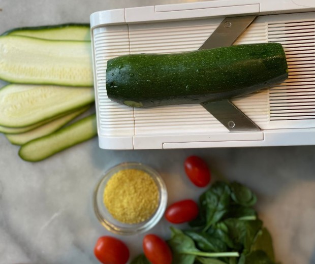 A zucchini on a mandoline slicer with thin slices beside it. Below are cherry tomatoes, spinach leaves, and a small bowl of yellow nutritional yeast on a light surface.
