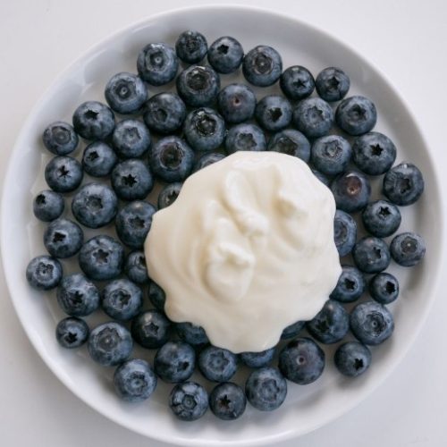 A white plate filled with fresh blueberries, with a generous dollop of creamy white yogurt placed in the center. The plate is set against a plain white background.