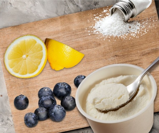 A wooden board with a sliced lemon, fresh blueberries, spilled salt from a shaker, and a bowl of white creamy yogurt with a spoon. The background is a gray marble surface.