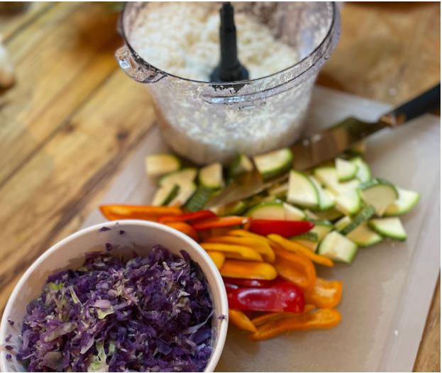 A cutting board with sliced yellow and red bell peppers, zucchini, and a knife sits beside a food processor with chopped cauliflower and a bowl of shredded purple cabbage.