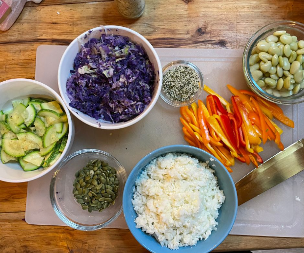 A wooden table with bowls of shredded purple cabbage, sliced cucumbers, white rice, lima beans, pumpkin seeds, hemp seeds, and sliced yellow and red bell peppers on a cutting board.
