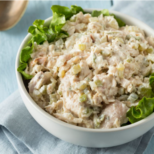 A white bowl of creamy chicken salad with chopped celery, served on a bed of green lettuce leaves, placed on a light blue cloth.