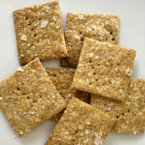 Seven square crackers sprinkled with sea salt are arranged on a white surface. The crackers have a golden-brown color and small holes evenly distributed across their surfaces.