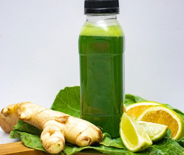 A bottle of green juice sits on leafy greens, surrounded by fresh ginger root, a slice of orange, and a wedge of lime, all arranged on a wooden surface with a plain background.