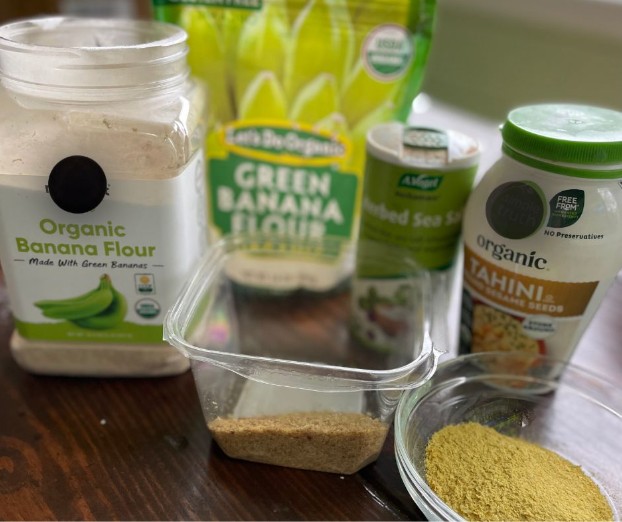 Various organic ingredients are displayed on a table, including banana flour, green banana flour, tahini, seeded sea salt, and a clear container with ground powder. The focus is on the product packaging and contents.