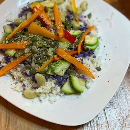 A white plate with rice topped with colorful vegetables, including carrots, zucchini, red pepper, and purple cabbage. The dish is garnished with seeds and served on a wooden surface.