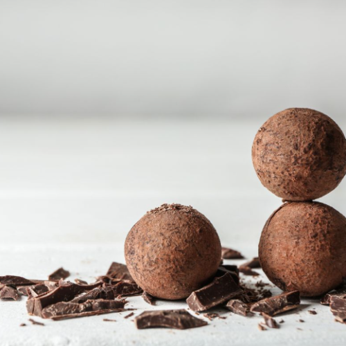 Three chocolate truffles dusted with cocoa powder, two stacked on top of each other, sitting on a white surface surrounded by pieces of broken dark chocolate.