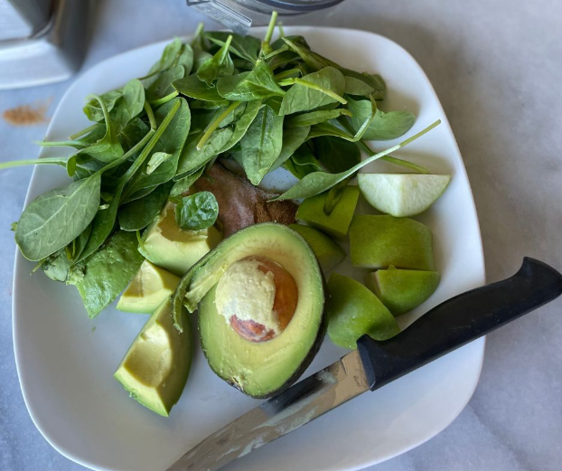 A white plate with fresh spinach, sliced green apple, halved avocado with pit, and knife on the side, all set on a marble surface.