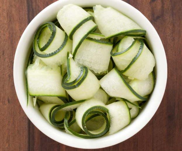 A white bowl filled with thinly sliced cucumber ribbons, some curled and arranged in a decorative pattern, placed on a wooden surface.