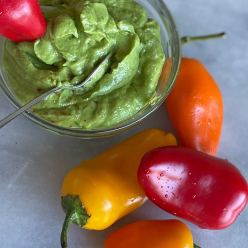 A glass bowl of creamy green guacamole with a spoon, next to fresh red, orange, and yellow mini bell peppers on a light gray surface.