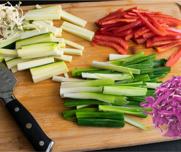 A wooden cutting board with sliced zucchini, red bell pepper, green onions, shredded purple cabbage, and a chef's knife on the left side.