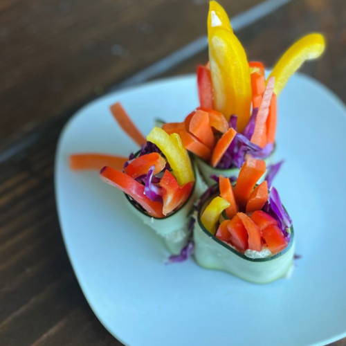 Three cucumber rolls filled with colorful sliced bell peppers and shredded red cabbage are arranged on a white plate atop a wooden surface. The vibrant vegetables are artfully displayed, creating a fresh, healthy presentation.