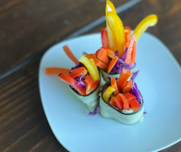 Three cucumber rolls filled with colorful sliced bell peppers and shredded red cabbage are arranged on a white plate atop a wooden surface. The vibrant vegetables are artfully displayed, creating a fresh, healthy presentation.