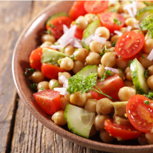 A bowl of chickpea salad with cherry tomatoes, cucumber slices, chopped red onion, and fresh parsley on a rustic wooden table.