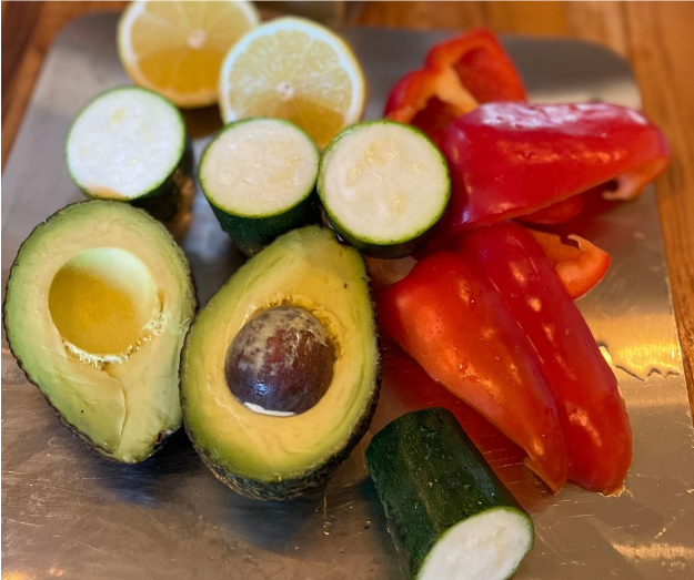 Avocado halves, sliced zucchini, red bell pepper pieces, and halved lemons are arranged on a metal surface with a wooden table in the background.