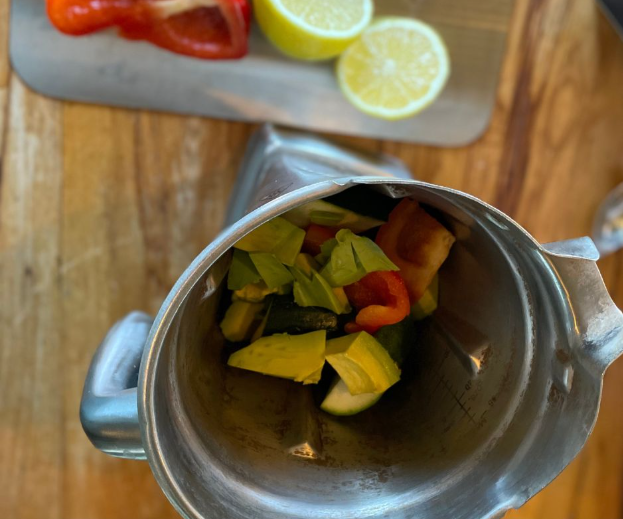 A blender containing chopped avocado, cucumber, and red bell pepper sits on a wooden counter. In the background, a cutting board holds sliced lemon halves and part of a red bell pepper.