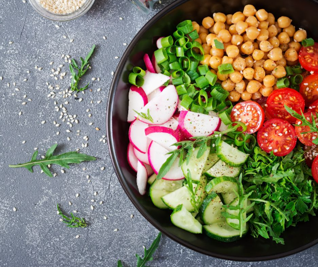 A black bowl filled with sliced radishes, cucumbers, cherry tomatoes, chickpeas, chopped green onions, fresh arugula, and parsley, with sesame seeds sprinkled on the gray surface nearby.