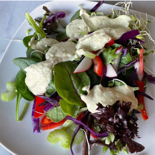 A plate of fresh mixed salad greens, red bell peppers, radishes, celery, and sprouts, topped with a creamy light-colored dressing, served on a white square plate.