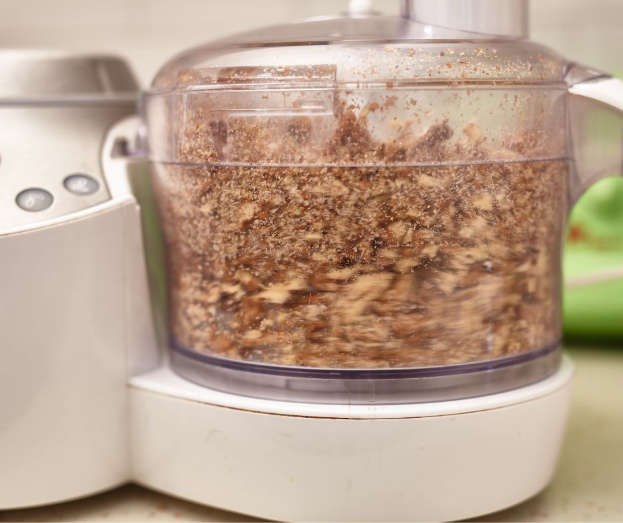 A close-up of a food processor blending nuts into a coarse mixture, with the inside container showing brown and tan nut pieces. The appliance is on a kitchen countertop.