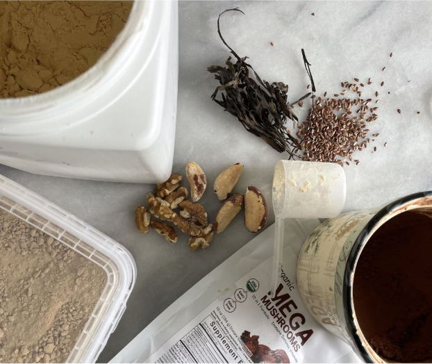 A flat lay of containers with powders, a scoop, walnuts, Brazil nuts, seaweed, flaxseeds, and a packet labeled “Mega Mushrooms” on a white marble surface.