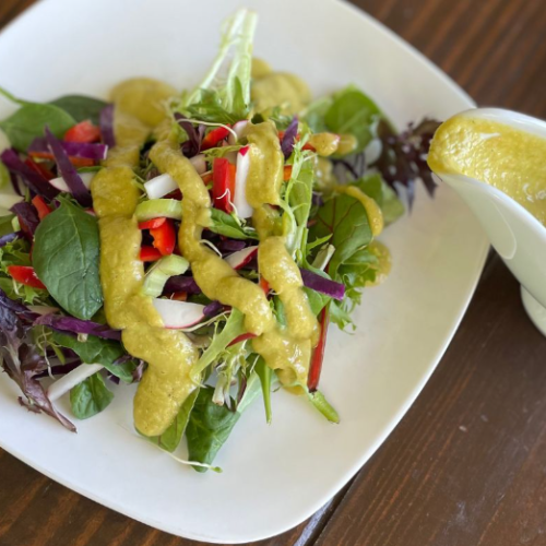 A fresh mixed green salad with red bell peppers and purple cabbage, topped with a generous drizzle of green dressing, is served on a white plate. A small gravy boat filled with more green dressing sits beside it.