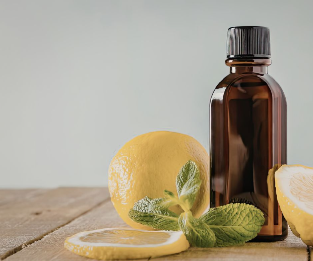 A brown glass bottle sits on a wooden surface next to a whole lemon, lemon slices, and a sprig of fresh mint. The background is light and blurred.