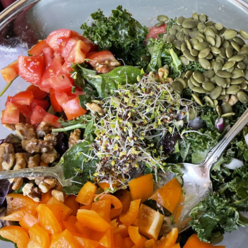 A glass bowl filled with a colorful salad containing chopped tomatoes, pumpkin seeds, kale, red onion, walnuts, yellow bell peppers, and a pile of sprouts, with two serving spoons inside.