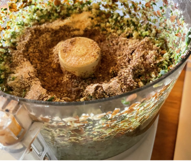 A close-up of a food processor containing finely chopped vegetables and various spices or seasonings, ready to be blended further.