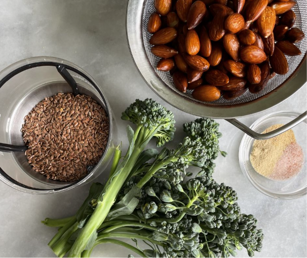 A bunch of fresh broccolini, a bowl of whole flaxseeds, a metal strainer with soaked almonds, and a small glass dish containing powdered seasonings on a gray surface.