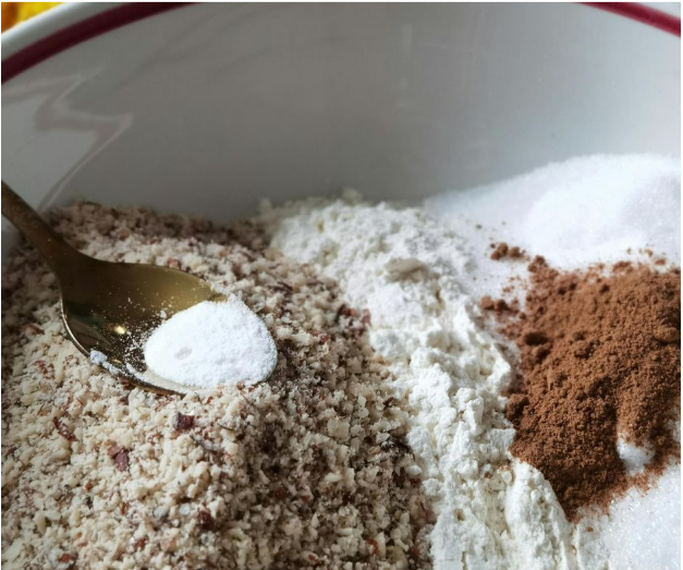 A close-up of a mixing bowl with ground nuts, flour, sugar, and cinnamon. A spoon is holding baking powder above the ground nuts, ready to be added to the ingredients.
