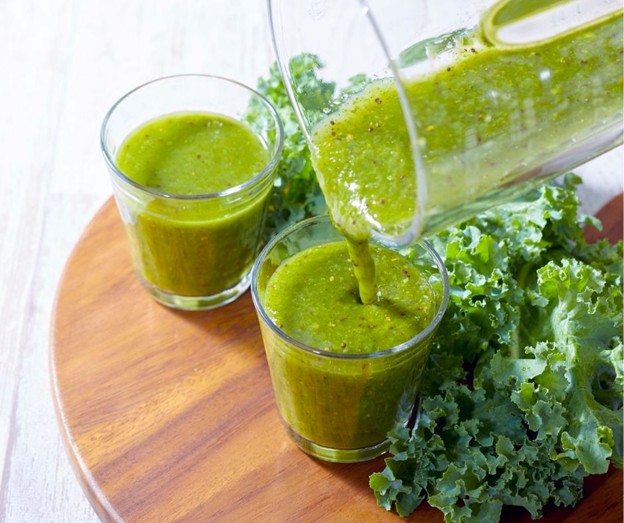 A glass pitcher pours a green smoothie into a clear glass beside another filled glass, both on a wooden tray, with fresh kale leaves arranged next to them.